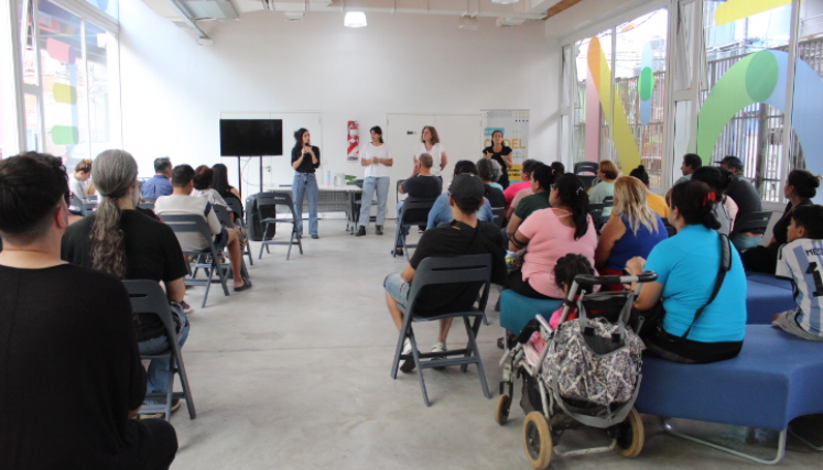 Personas sentadas en un salón amplio y luminoso escuchan a un grupo de expositoras al frente durante una charla comunitaria con participación de familias