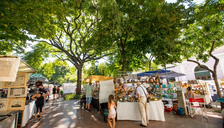 Vista general de la Feria de San Telmo en la Plaza Dorrego. Bajo la sombra de grandes árboles, se observan puestos con artesanías, antigüedades y personas recorriendo el paseo.