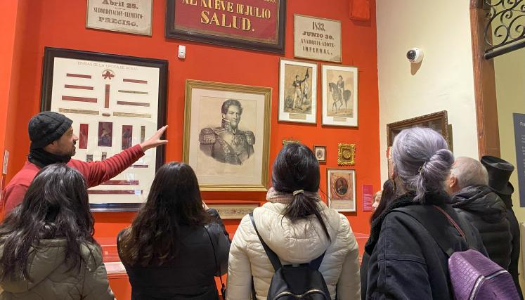Imagen de un guía del museo señalando objetos sobre el rosismo. De espaldas se observan varias personas adultas prestando atención