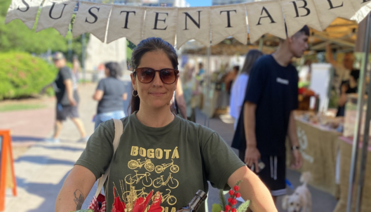 Mujer con bandeja con objetos en venta. De fondo feria con cartel que dice Sustentable